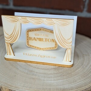 Peut inclure: Une sc&egrave;ne en bois avec une enseigne de cin&eacute;ma qui indique "Now Playing Hamilton". La sc&egrave;ne est d&eacute;cor&eacute;e de rideaux et repose sur une base en bois.