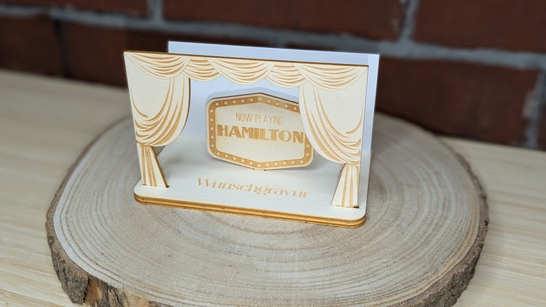 Peut inclure: Une sc&egrave;ne en bois avec une enseigne de cin&eacute;ma qui indique "Now Playing Hamilton". La sc&egrave;ne est d&eacute;cor&eacute;e de rideaux et repose sur une base en bois.