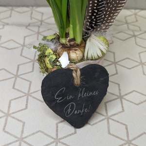 May include: A small potted plant with green leaves and a heart-shaped slate sign that says "Ein kleines Danke!" The plant is sitting on a white and brown patterned surface.