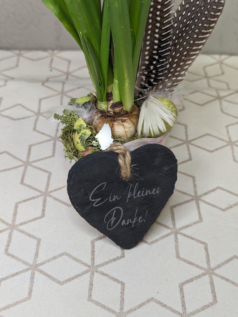 May include: A small potted plant with green leaves and a heart-shaped slate sign that says "Ein kleines Danke!" The plant is sitting on a white and brown patterned surface.