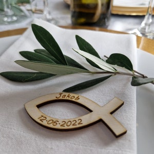 Könnte beinhalten: Eine hölzerne Fisch-Platzkarte mit dem Namen "Jakob" und dem Datum "12-06-2022" eingraviert. Die Platzkarte liegt auf einer weißen Serviette mit einem grünen Olivenzweig.