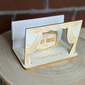 Peut inclure: Un porte-cartes en bois avec une d&eacute;coupe en forme de rideau de sc&egrave;ne et une pancarte indiquant "It's on King Time". Le porte-cartes est pos&eacute; sur une surface en bois.