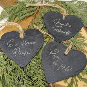 May include: Three black heart-shaped slate ornaments with twine hangers. The ornaments are engraved with the text "Familie Schmidt", "Ein kleines Danke!" and "Papa, du fehlst".