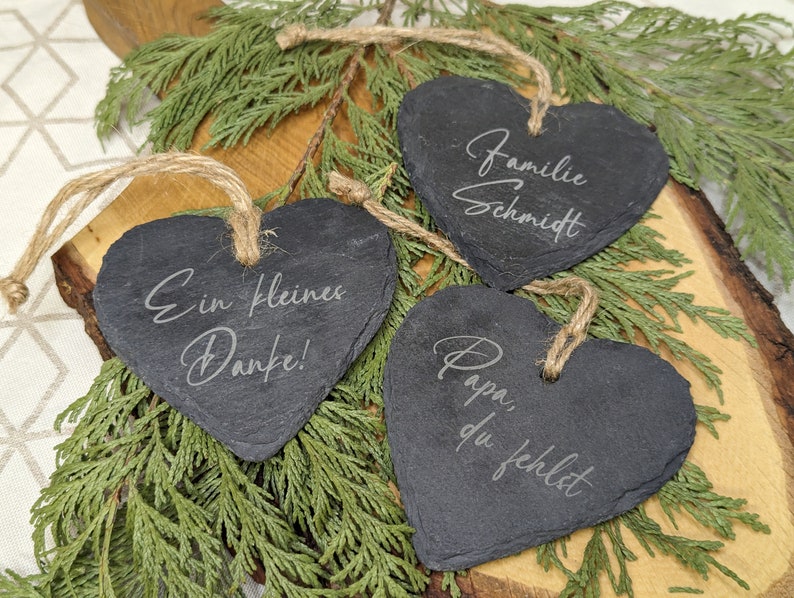 May include: Three black heart-shaped slate ornaments with twine hangers. The ornaments are engraved with the text "Familie Schmidt", "Ein kleines Danke!" and "Papa, du fehlst".