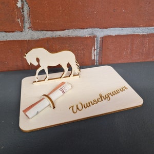 Op de afbeelding: Een houten paardenfiguur met een gleuf voor geld. Het paard staat op een houten voet met de tekst "Wunschgrauwun" erop gegraveerd.