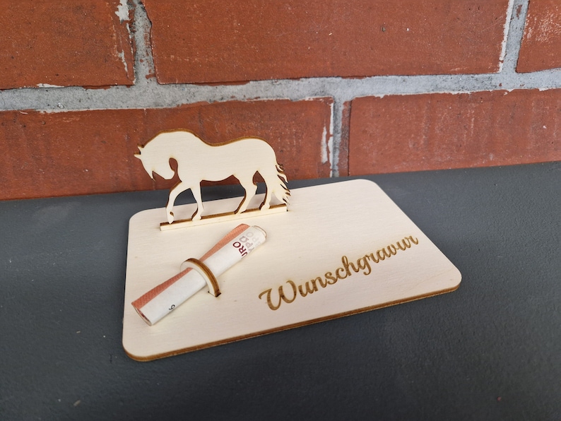 Op de afbeelding: Een houten paardenfiguur met een gleuf voor geld. Het paard staat op een houten voet met de tekst "Wunschgrauwun" erop gegraveerd.