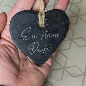 May include: A black heart-shaped slate sign with the text "Ein kleines Danke!" written in white. The sign is hanging from a twine string.