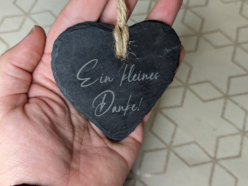 May include: A black heart-shaped slate sign with the text "Ein kleines Danke!" written in white. The sign is hanging from a twine string.