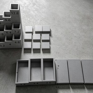 May include: A light grey concrete desk organiser set featuring a tiered block with nine square compartments labelled 3.2, 4, 4.8, 5.6, 6.4, 7.2, 8, 9.6, and 12.8, alongside nine square tiles and three rectangular trays labelled 3.8, 3.4, and 3.6.