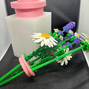 May include: A pink, tiered, cylindrical container sits on a white, square base. A bouquet of green-stemmed, white daisy and purple lavender flowers is arranged in a pink vase. The flowers have yellow centres.