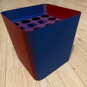May include: A square-shaped container with a gradient finish transitioning from deep blue to red. The container features a flat interior base with a grid of circular cutouts, and the exterior walls are smooth with visible horizontal stacking lines.
