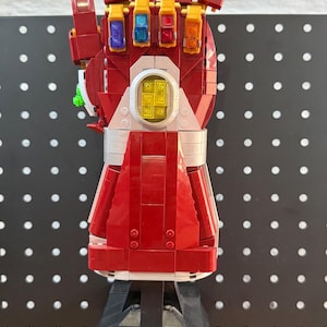Op de afbeelding: Een rood en goudkleurig Nano Gauntlet-model gemaakt van Lego-stenen. De handschoen heeft zes gekleurde edelstenen en is gemonteerd op een zwarte basis met de tekst "LEGO INFINITY SAGA" en "NANO GAUNTLET".