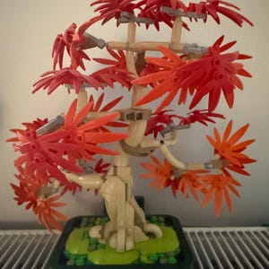 Puede incluir: Un modelo de árbol bonsái construido con ladrillos de plástico. El árbol presenta un tronco y ramas beige, con vibrantes racimos de hojas rojas y naranjas. Está colocado en una maceta rectangular verde oscuro con base negra.
