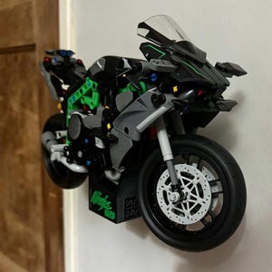 May include: A black, gray, and green building block motorcycle model featuring a clear windshield, black tires, and a display stand with the word Ninja printed in green. The structure includes visible mechanical components and intricate frame details.