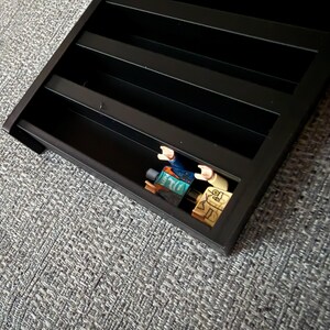 May include: A black display shelf with multiple horizontal tiers designed for organising small items. One compartment contains a small plastic figure with a blue torso, tan arms, and a yellow head with printed facial features and glasses.