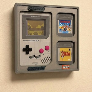 Può includere: Un espositore grigio per Game Boy con una console Game Boy e due cartucce di gioco. La console ha uno schermo che mostra una scena desertica, un pad direzionale nero e pulsanti rosa. Le cartucce sono etichettate "Super Mario Land" e "Zelda".