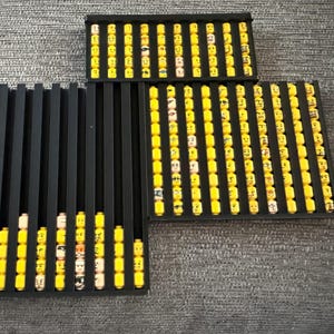 May include: Black trays filled with yellow Lego heads with various facial expressions. The trays are arranged on a gray surface. The Lego heads are neatly organized, showcasing a collection of minifigure parts.