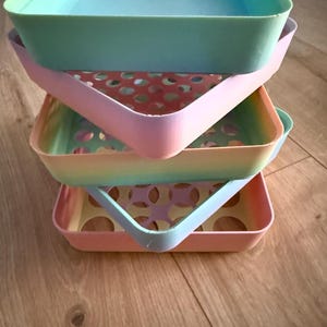 May include: A stack of four square plastic storage trays in pastel colours including mint green, soft pink, yellow, and light blue. Each tray features a perforated base with circular cutouts and smooth, rounded corners, arranged in a vertical column.