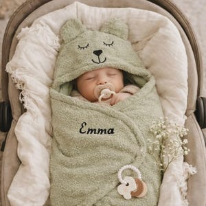 May include: A sleeping baby wrapped in a soft, green bear-themed hooded towel. The towel is embroidered with the name "Emma". A pacifier is in the baby's mouth, and a wooden teether is attached to the towel. The baby is in a grey car seat.