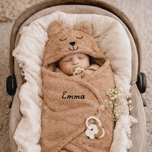 Op de afbeelding: Een slapende baby gewikkeld in een bruine deken met berenmotief, met de naam "Emma" geborduurd op de deken. De baby ligt in een autostoeltje, met een speen en een houten bijtring.