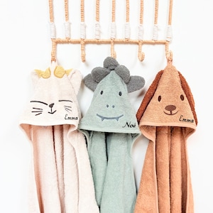 Op de afbeelding: Drie badhanddoeken met capuchon voor baby's hangen aan een decoratief rek. De handdoeken zijn lichtroze, mintgroen en bruin, elk met een ander dierengezicht: een kat, een dinosaurus en een hond. Elke handdoek heeft een geborduurde naam.