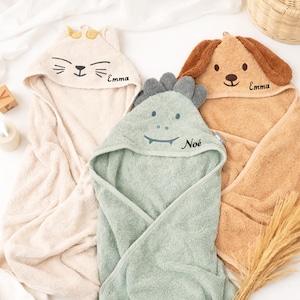 May include: Three hooded baby towels in beige, sage green, and tan. The towels are shaped like a cat, dinosaur, and dog, each with embroidered details and a name. The cat and dog towels have the name "Emma", and the dinosaur towel has the name "Noé".