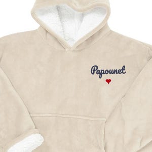 May include: A beige hooded blanket with a white sherpa lining in the hood and cuffs. The word "Papounet" is embroidered in navy blue script with a small red heart below it. The blanket has a large front pocket.