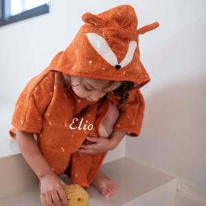 May include: A child wearing an orange hooded towel with a fox face on the hood. The towel has the name "Elio" embroidered on the front.