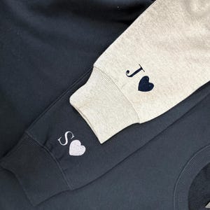 May include: Two sweatshirts, one navy blue and one light grey, with embroidered initials and heart symbols. The navy blue sweatshirt has the initial "S" and a white heart on the sleeve. The light grey sweatshirt has the initial "J" and a navy blue heart on the sleeve.
