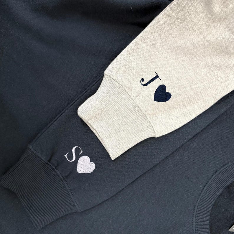 May include: Two sweatshirts, one navy blue and one light grey, with embroidered initials and heart symbols. The navy blue sweatshirt has the initial "S" and a white heart on the sleeve. The light grey sweatshirt has the initial "J" and a navy blue heart on the sleeve.