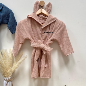 Könnte beinhalten: Ein rosa Bademantel mit Kapuze, Hasenohren, einem Bindegürtel und dem Namen "Emma" auf der Vorderseite bestickt.