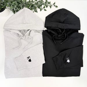 May include: Two hooded sweatshirts, one light grey and one black, are displayed. The grey sweatshirt has the letter "K" and a heart embroidered on the cuff. The black sweatshirt has the letter "S" and a heart embroidered on the cuff.