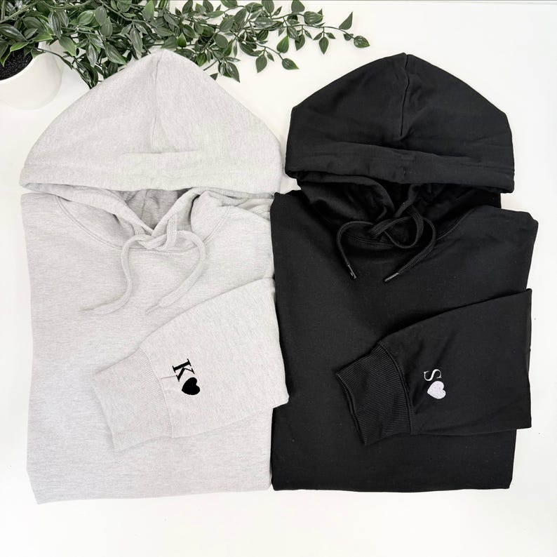 May include: Two hooded sweatshirts, one light grey and one black, are displayed. The grey sweatshirt has the letter "K" and a heart embroidered on the cuff. The black sweatshirt has the letter "S" and a heart embroidered on the cuff.