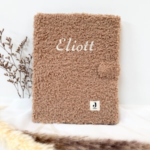 May include: A brown fuzzy notebook with the name "Eliott" embroidered on the front. The notebook has a small white label with a "J" on it.