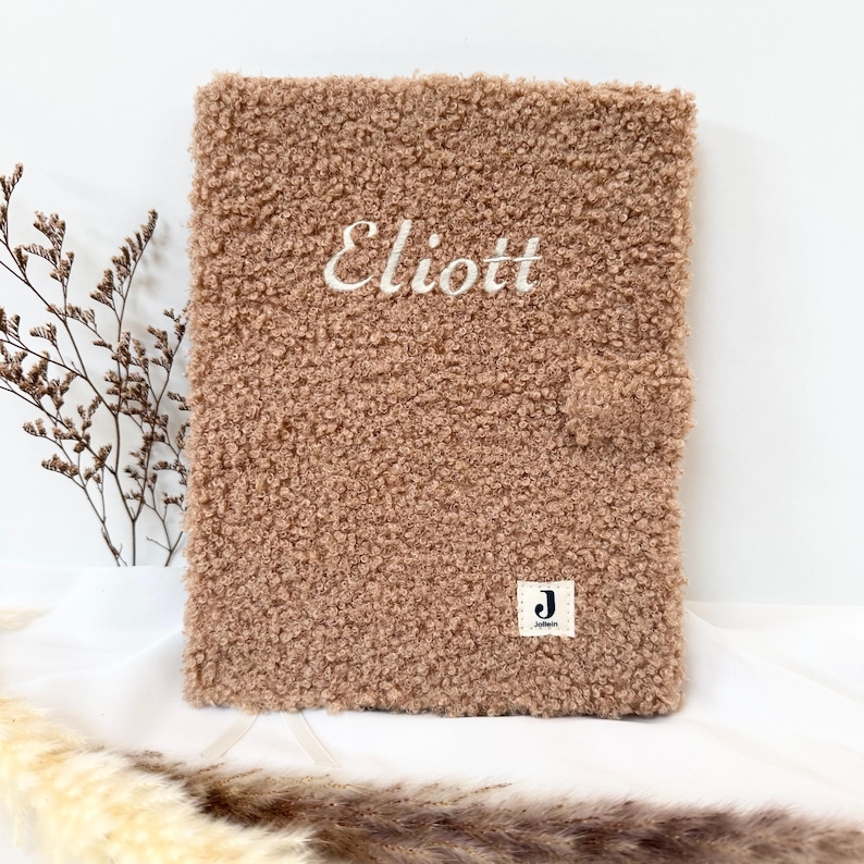 May include: A brown fuzzy notebook with the name "Eliott" embroidered on the front. The notebook has a small white label with a "J" on it.