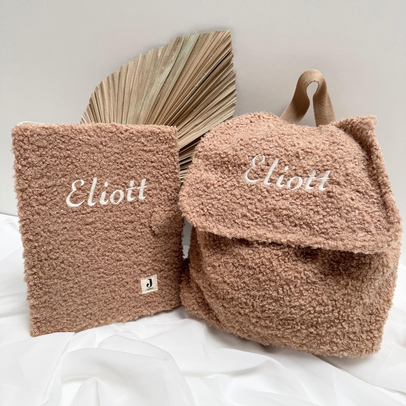 May include: Two brown fuzzy bags with the name "Eliott" embroidered on them. The larger bag is a backpack with a brown strap. The smaller bag is a flat pouch.