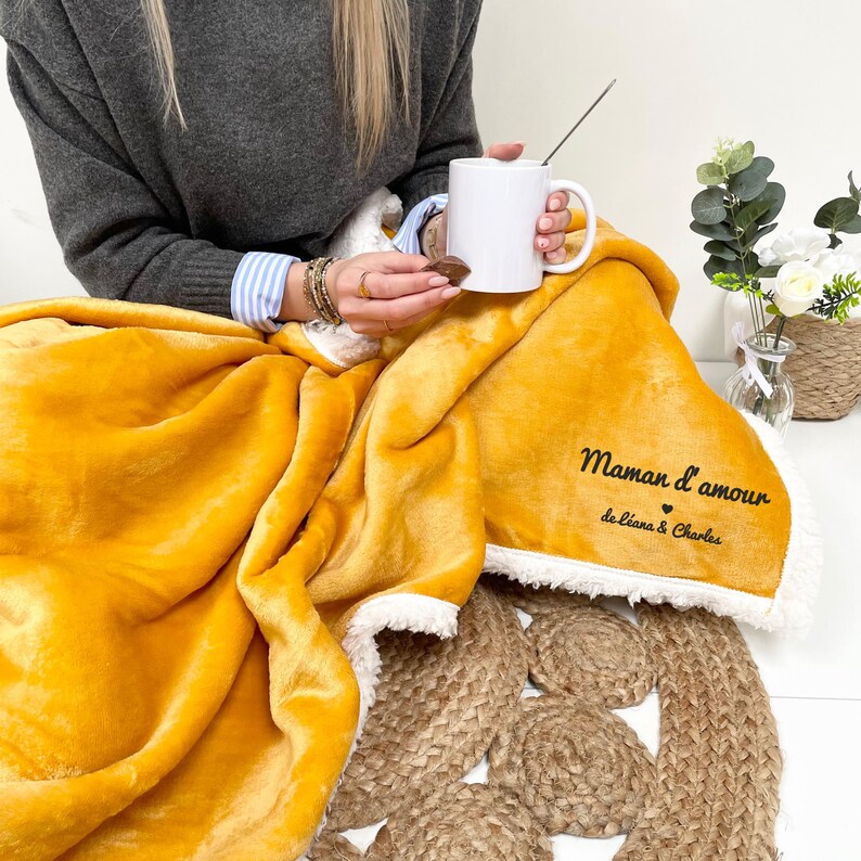 May include: A mustard yellow fleece blanket with a white sherpa trim. The blanket has the text "Maman d'amour de Eliana & Charles" embroidered on it.
