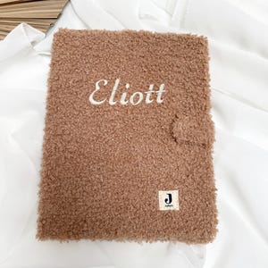 May include: A brown fuzzy notebook with the name "Eliott" embroidered on the front. The notebook has a small white tag with the letter "J" and the word "Jolie" on it.