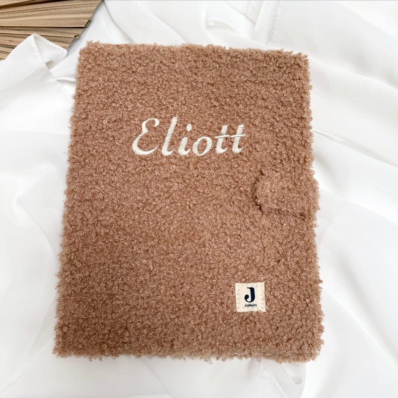 May include: A brown fuzzy notebook with the name "Eliott" embroidered on the front. The notebook has a small white tag with the letter "J" and the word "Jolie" on it.