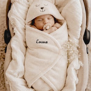 May include: A sleeping newborn baby wrapped in a cream towel and bear-eared hood, in a car seat. The baby has a dummy. The name "Emma" is embroidered on the towel. A sprig of white flowers is placed nearby.