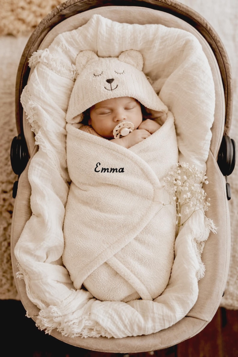 May include: A sleeping newborn baby wrapped in a cream towel and bear-eared hood, in a car seat. The baby has a dummy. The name "Emma" is embroidered on the towel. A sprig of white flowers is placed nearby.