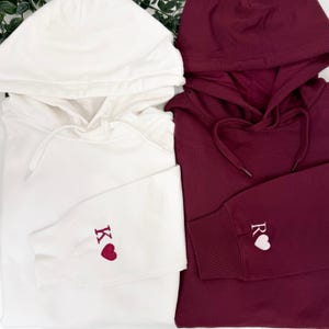 May include: Two hooded sweatshirts, one white and one burgundy. The white sweatshirt has the letter "K" and a heart embroidered on the sleeve. The burgundy sweatshirt has the letter "R" and a heart embroidered on the sleeve.