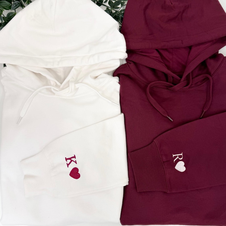 May include: Two hooded sweatshirts, one white and one burgundy. The white sweatshirt has the letter "K" and a heart embroidered on the sleeve. The burgundy sweatshirt has the letter "R" and a heart embroidered on the sleeve.