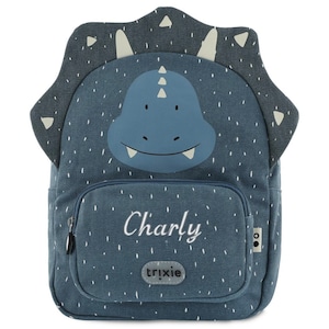 May include: A blue dinosaur-themed backpack with the name "Charly" embroidered on the front pocket. The backpack features a dinosaur head design on the top and a small front pocket with a zipper. The brand "trixie" is also visible.
