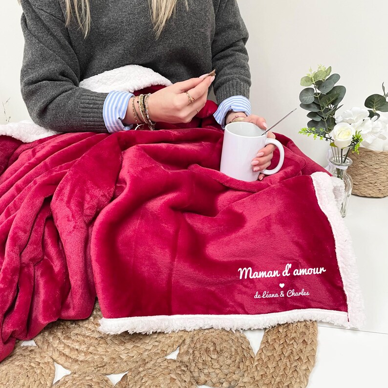 May include: A red fleece blanket with a white sherpa lining. The blanket has the text "Maman d'amour de Liana & Charles" embroidered on it.