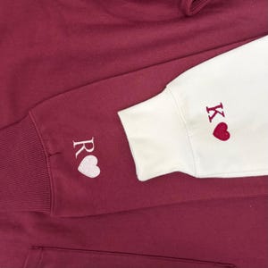 May include: Burgundy hoodie with white and burgundy embroidered details. The left sleeve cuff features the letter "R" and a white heart. The right sleeve cuff is white with the letter "K" and a red heart. The hoodie has a drawstring hood.