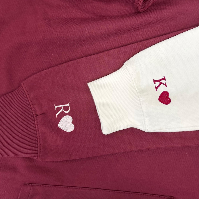 May include: Burgundy hoodie with white and burgundy embroidered details. The left sleeve cuff features the letter "R" and a white heart. The right sleeve cuff is white with the letter "K" and a red heart. The hoodie has a drawstring hood.