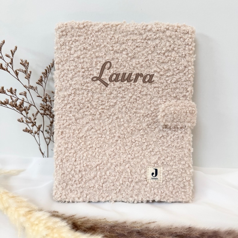 May include: A light beige fuzzy notebook with the name "Laura" embroidered on the front. The notebook has a small brown label with the letter "J" on it.