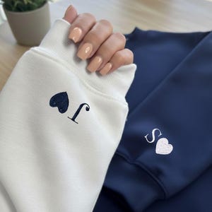 May include: A close-up of two sweatshirts, one white and one navy blue. The white sweatshirt has a dark blue heart and the letter "J" embroidered on the cuff. The navy blue sweatshirt has a white heart and the letter "S" embroidered on the cuff.
