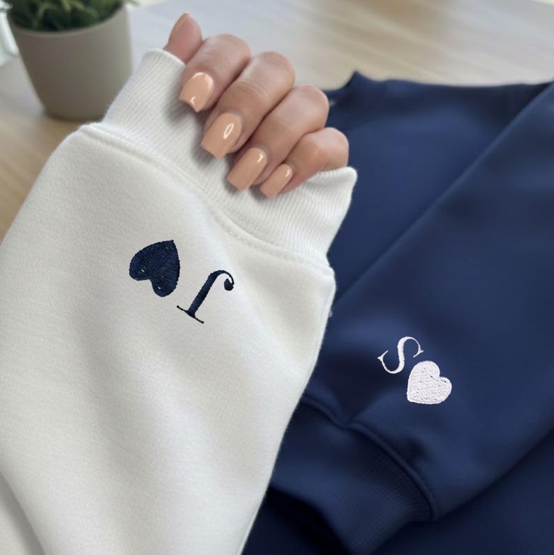 May include: A close-up of two sweatshirts, one white and one navy blue. The white sweatshirt has a dark blue heart and the letter "J" embroidered on the cuff. The navy blue sweatshirt has a white heart and the letter "S" embroidered on the cuff.